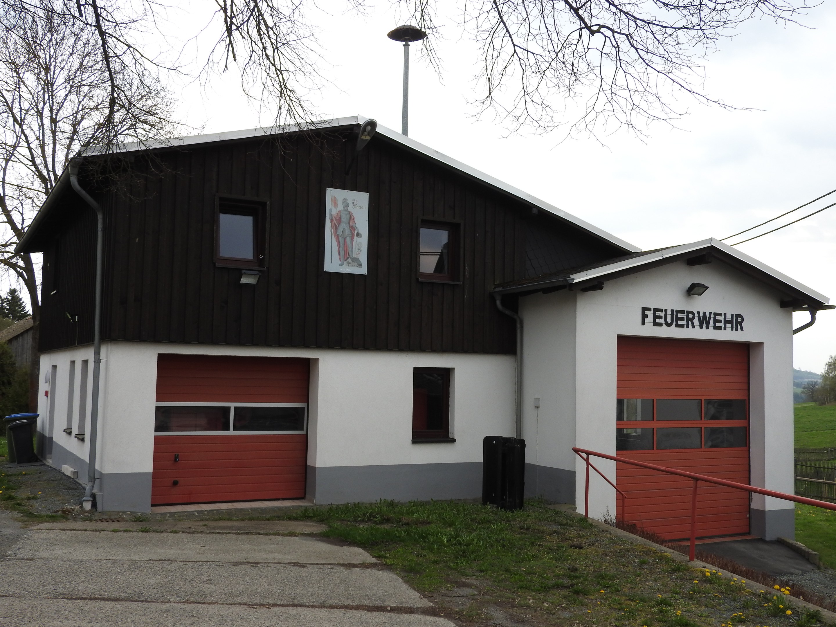 Rote und weiße Feuerwache-Gebäude neben einer Straße mit Gras, gelben Blumen und Bäumen, unter einem Himmel-Hintergrund.
