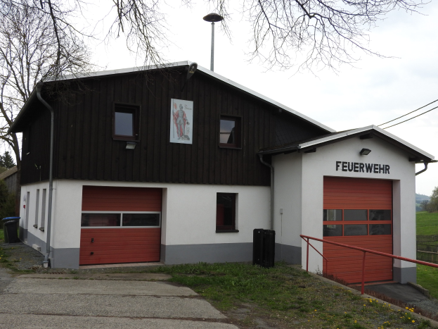 Rote und weiße Feuerwache-Gebäude neben einer Straße mit Gras, gelben Blumen und Bäumen, unter einem Himmel-Hintergrund.