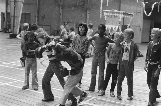 Eine Gruppe von Kindern, die auf einem Basketballfeld spielt, mit zwei kämpfenden Personen in der Mitte und anderen auf den Tribünen im Hintergrund, dargestellt in Schwarz-Weiß.