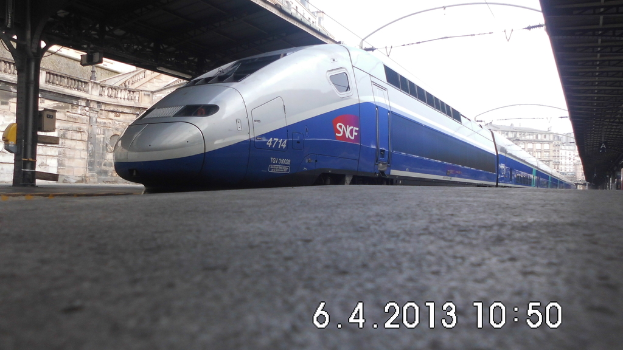 Ein Zug ist in der Mitte des Bildes mit einem Bahnsteig und Text unten, Gebäuden und einem Dach links und Gebäuden, Dächern, Kabeln und Himmel oben.