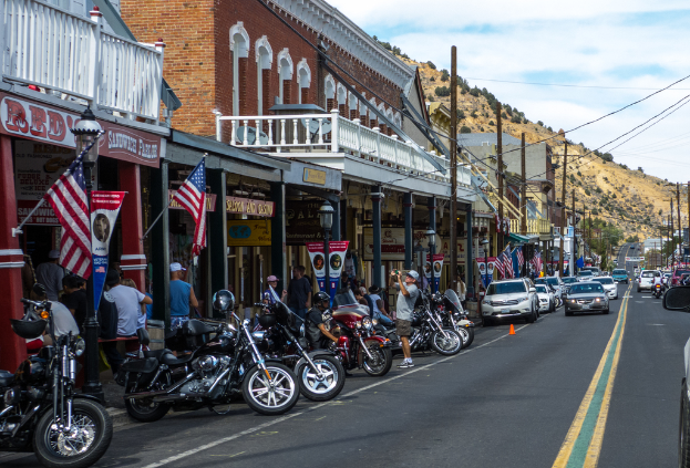 Fahrräder und Autos stehen auf einer Straße mit Menschen in der Nähe, Gebäude mit Flaggen im Hintergrund, ein Hügel mit Bäumen und einen klaren Himmel darüber.