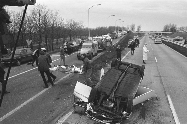 Schwarzes und weißes Bild eines umgestürzten Autos am Straßenrand mit beschädigten Fahrzeugen in der Nähe und einer Gruppe von Menschen drumherum, mit Laternen, Bäumen, einer Brücke und Himmel im Hintergrund.