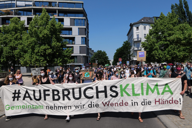 Menschen mit Masken halten ein Banner mit der Aufschrift 'Aufbruchsklima' vor einem Gebäude mit Bäumen und einem klaren Himmel.