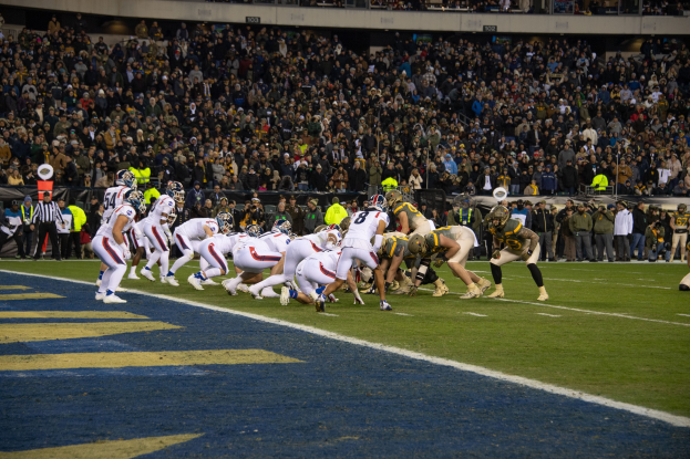 Ein Fußballspiel im Gange mit Spielern in Sportkleidung und Helmen auf dem Boden, umgeben von Tribünen, Geländern und Werbetafeln.