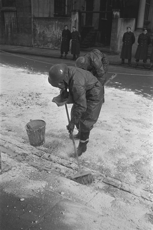 Ein Mann in einem Helm und Handschuhen kehrt den Schnee vom Gehweg mit einer Besen, während eine Gruppe von Menschen auf dem Bürgersteig in der Nähe steht, mit einem Gebäude mit Fenstern und einer Tür im Hintergrund.