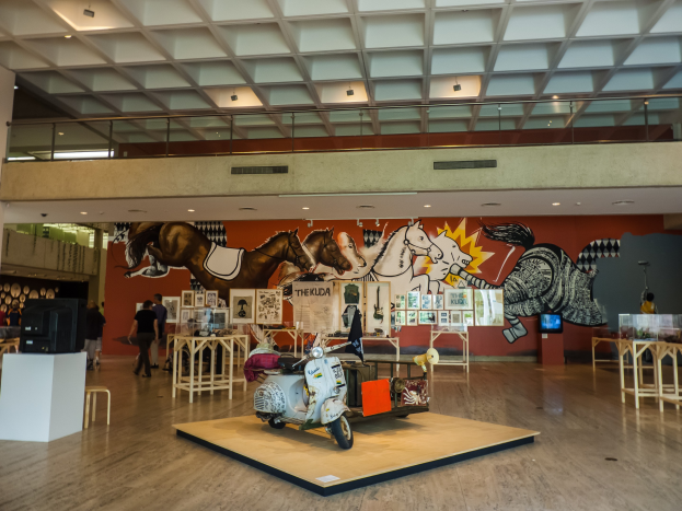 Ein großer Ausstellungsraum im Nationalen Kunstmuseum mit Tischen, Stühlen, einem Motorroller, einem Fernseher, stehenden Besuchern, Foto Rahmen, einer Wandmalerei und Deckenleuchten.