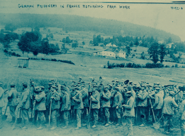 Eine Gruppe deutscher Gefangener in Frankreich, die Gewehre halten und auf dem Boden stehen, mit Bäumen, Häusern und einem klaren blauen Himmel im Hintergrund.