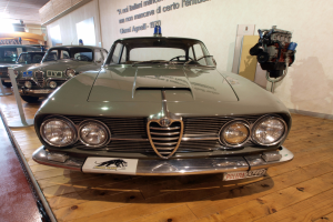 Ein klassischer Alfa Romeo Giulietta Sprint GT Junior aus den 1970er Jahren wird in einem Museum ausgestellt, abgesperrt durch Seilbarrieren mit Text an der Wand dahinter bei warmem Deckenlicht.