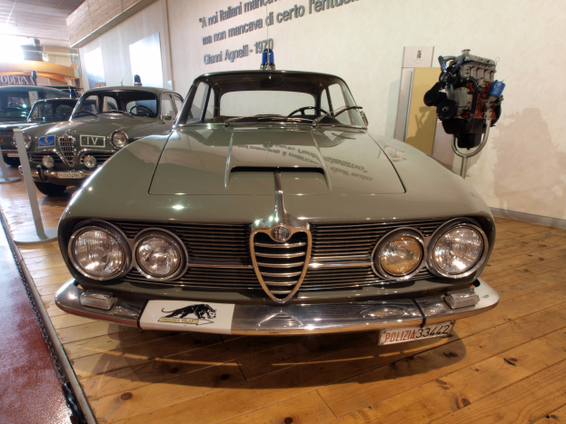 Ein klassischer Alfa Romeo Giulietta Sprint GT Junior aus den 1970er Jahren wird in einem Museum ausgestellt, abgesperrt durch Seilbarrieren mit Text an der Wand dahinter bei warmem Deckenlicht.