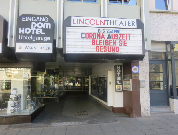 Außenansicht des Lincoln Theaters in Berlin mit Glasfenstern und -türen sowie einer Tafel mit Text und einer belebten Stadtlandschaft im Inneren.