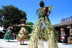 Drei Personen in Kostümen tanzen in der Mitte, mit Geschäften, die Kleidung und Gegenstände im Hintergrund anbieten, Text auf einem Gebäude, zusätzlichen Geschäften und Bäumen links, einem Weg am Boden und Himmel oben.