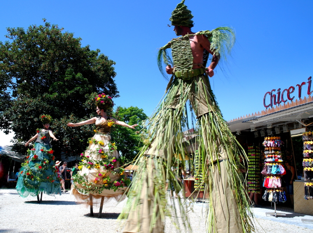 Drei Personen in Kostümen tanzen in der Mitte, mit Geschäften, die Kleidung und Gegenstände im Hintergrund anbieten, Text auf einem Gebäude, zusätzlichen Geschäften und Bäumen links, einem Weg am Boden und Himmel oben.