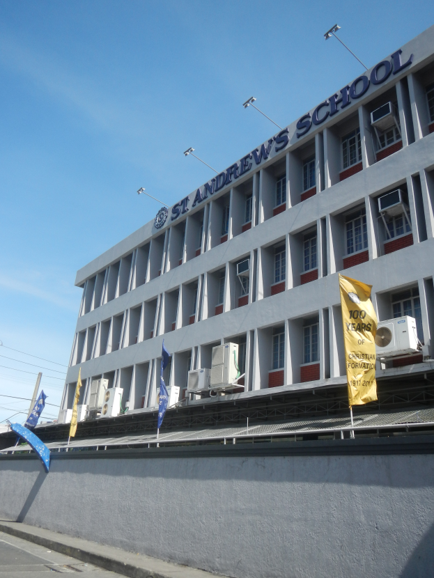 Ein Schulgebäude mit Fenstern, Klimaanlagen, Flaggen, einem Namenstafel, Straßenlaternen, Strommasten, Stromkabeln und einer bewölkten Himmel im Hintergrund.