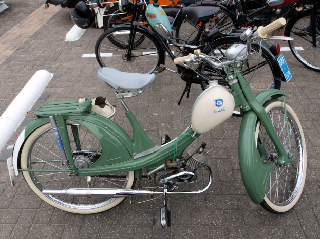 Ein grünes Moped mit weißem Rahmen und schwarzem Sitz, das auf einem Gehweg neben anderen Motorrädern geparkt ist, mit ein paar Menschen in der Nähe.