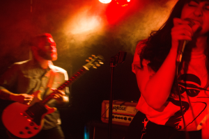 Eine Frau singt in ein Mikrofon auf der Bühne, während ein Mann Gitarre spielt und Deckenleuchten die lebendige Szenerie erhellen.