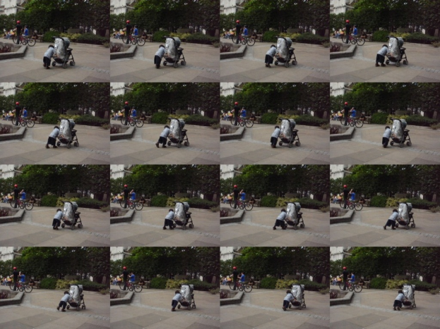 Ein Kind schiebt einen Wagen auf der Straße, während zwei Personen Fahrräder halten, Bäume im Hintergrund und ein Gebäude links im Bild.