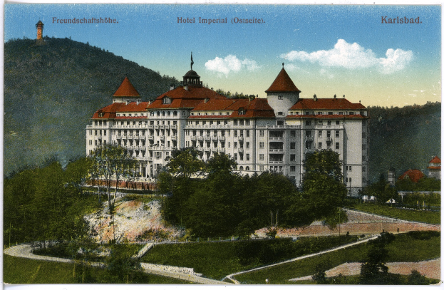 Eine alte Postkarte, die das Hotel Imperial in Karlsbad, Deutschland zeigt, ein großes Gebäude mit zahlreichen Fenstern, umgeben von Bäumen, Pflanzen und Gras, mit einem Weg im Vordergrund, Hügel im Hintergrund und einem bewölkten Himmel, mit Text oben auf dem Bild.