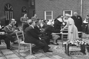 Ein Schwarz-Weiß-Foto einer Hochzeitszeremonie vor einem Gebäude, mit Sitzenden und Stehenden sowie einem Blumenstrauß im Vordergrund, das Gebäude hat Fenster und eine Leitung an der Wand.