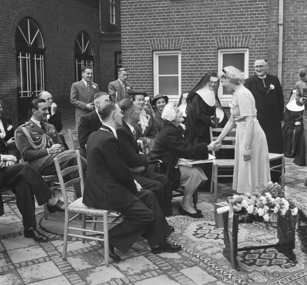 Ein Schwarz-Weiß-Foto einer Hochzeitszeremonie vor einem Gebäude, mit Sitzenden und Stehenden sowie einem Blumenstrauß im Vordergrund, das Gebäude hat Fenster und eine Leitung an der Wand.