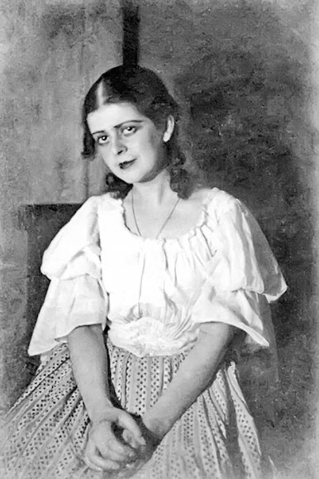 Ein junges Mädchen in einem weißen Kleid sitzt auf einem Stuhl vor einer Wand in einem alten Schwarz-Weiß-Foto, das angeblich Anna Pavlova ist.