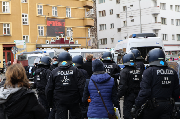 Polizeibeamte in Uniform vor einer Menge bei einer Demonstration in Berlin, mit Fahrzeugen, Gebäuden und einem Fotografen im Hintergrund.