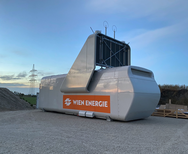 Eine große weiße und orangefarbene Windturbine steht auf einem Schmutzfeld umgeben von Bäumen und einem Turm im Hintergrund, mit ein paar Gegenständen rechts auf dem Boden und dem Himmel oben sichtbar.