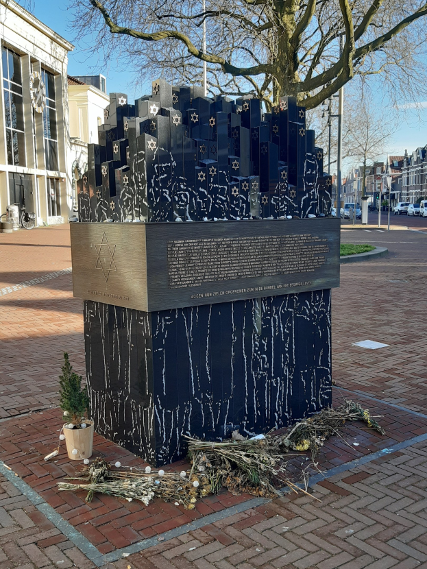 Denkmal in einem Stadtplatz für Holocaust-Opfer, mit einem Baum davor, einer Topfpflanze auf dem Boden, verstreuten trockenen Blättern und Gebäuden, Fahrzeugen, Masten und Bäumen im Hintergrund.