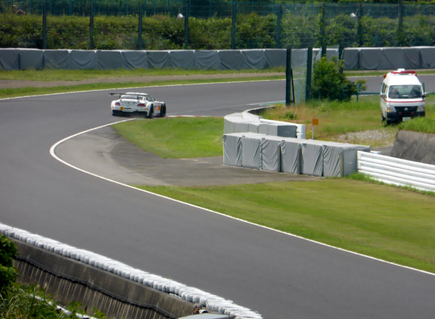 Ein Rennwagen fährt eine Strecke entlang, im Hintergrund ist ein Polizeiwagen zu sehen, umgeben von Gras, Pflanzen, Bäumen und Absperrungen, mit einem Fahrzeug auf der rechten Seite und Text am unteren Bildrand.