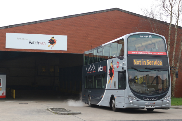 Doppeldeckerbus mit Rauchwolke an einem Gebäude vorbeifahrend, mit Gras, Bäumen und einem blauen Himmel im Hintergrund und einem sichtbaren Fahrzeugkennzeichen.