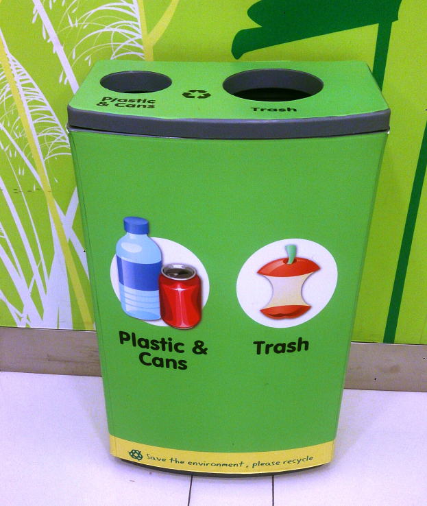 Ein grüner Recycling-Eimer, der mit Plastik und Dosen gefüllt ist, steht auf einem weißen Boden, mit einem Plakat im Hintergrund an der Wand.