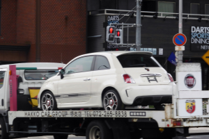 Ein weißes Auto wird von einem Abschleppwagen auf einer Straße mit Gebäuden, Masten, Verkehrszeichen und Schildern im Hintergrund geschleppt.