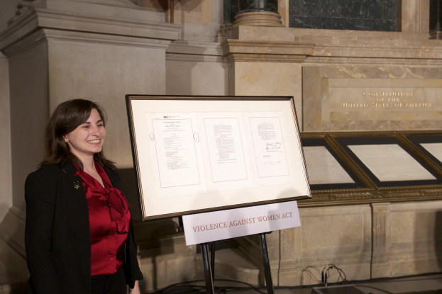 Eine Frau in einem roten Shirt und schwarzer Jacke lächelt vor einem Schild, auf dem "Gesetz gegen Gewalt an Frauen" steht, mit einer Wand, Säulen und anderen Gegenständen im Hintergrund.