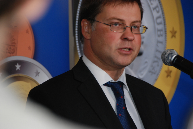 Ein Mann in einer schwarzen Jacke, weißem Hemd und blauer Krawatte spricht in ein Mikrofon vor einem blauen Plakat mit Logos.