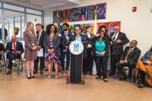 Gruppe von Menschen vor einem Rednerpult mit Mikrofon und einer Tafel mit Text, einige sitzen auf Stühlen und andere stehen, mit Fahnen, einer Tafel, einem Feuerlöscher und Deckenleuchten an der Wand.