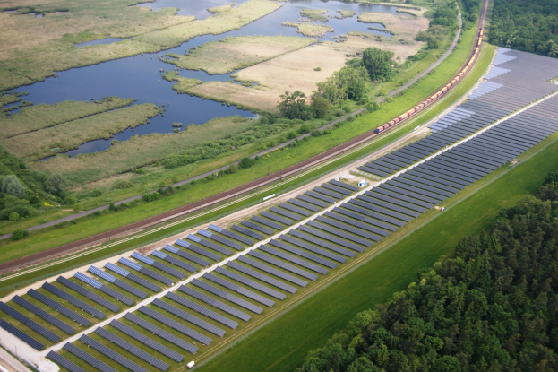 Luftbild eines Solarparks mit Panelen, umgeben von Bäumen, Gras, Wasser und einer nahen Bahnstrecke.