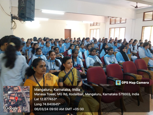 Eine Gruppe von Menschen sitzt in Stühlen in einem Raum mit einer Wand, Fenstern auf der rechten Seite, Deckenleuchten und einem Ventilator und einem Redner auf der linken Seite, mit Text und einer Karte unten.