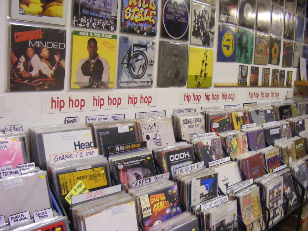 Ein Metallständer mit Büchern und optischen Disc-Cases, jeweils verziert mit Postern mit Text und Bildern, sowie zusätzlichem Text auf Papieren, die an der Wand in der Mitte angebracht sind.