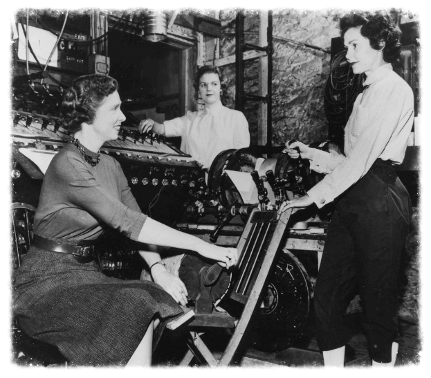 Drei Frauen bei der Arbeit an einer Maschine in einer Fabrik, wobei eine in der Mitte sitzt und strahlt und zwei auf jeder Seite stehen und die Maschine halten, in Schwarz-Weiß.