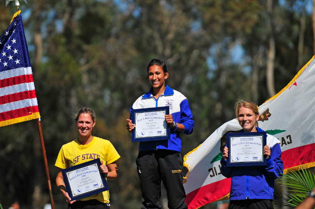 Drei Frauen mit Zertifikaten in der Hand, mit Flaggen und Bäumen im Hintergrund.