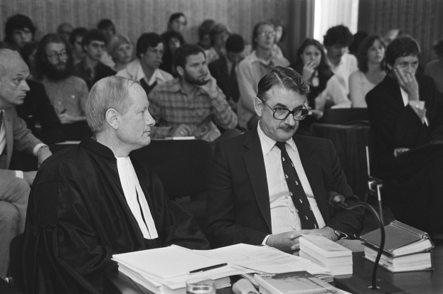 Oberster Gerichtshof Richter John Roberts in einer schwarzen Robe und ein anderer Mann in einem Anzug an einem Tisch mit Büchern, Papieren, einem Glas und einem Mikrofon, vor einer Menge mit einer Wand und einem Vorhang im Hintergrund.