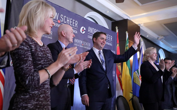 Eine Gruppe von Menschen, die vor Fahnen stehen und klatschen, mit einem Tisch mit Gegenständen rechts daneben, Stühlen, einer Fahne und einer Wand im Hintergrund und Lampen und einem Ventilator auf dem Dach.