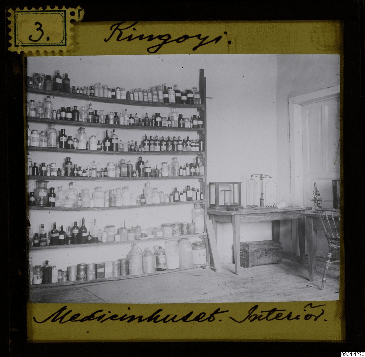 Schwarze und weiße Fotografie eines alten Apothekeninneren mit Regalen voller Flaschen, einem Tisch mit Gegenständen, einem Stuhl, einer Tür und einer Wand, mit Text unten.