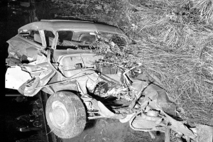 Ein Schwarz-Weiß-Foto eines schwer beschädigten Autos am Straßenrand mit einer zerbeulten Front, zersplitterten Fenstern, einem Baum im Hintergrund und verstreuten Gegenständen rund um die Unfallstelle.