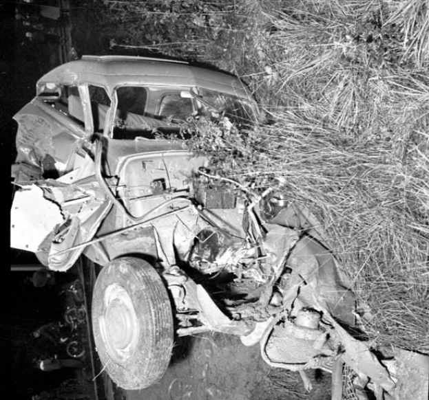 Ein Schwarz-Weiß-Foto eines schwer beschädigten Autos am Straßenrand mit einer zerbeulten Front, zersplitterten Fenstern, einem Baum im Hintergrund und verstreuten Gegenständen rund um die Unfallstelle.