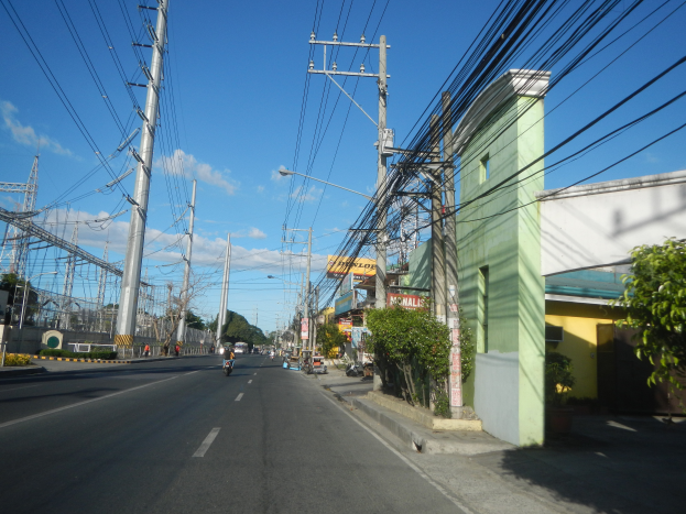 Eine Stadtstraße mit Fahrzeugen, Stromleitungen, Strommasten, Gebäuden, Bäumen, Pflanzen, Texttafeln, Laternen und Türmen unter einem bewölkten Himmel.