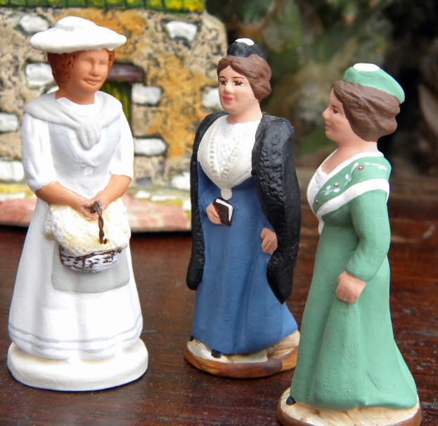 Drei geschnitzte Puppen sind auf einer Holzplatte mit Spielzeughäusern im Hintergrund angeordnet.