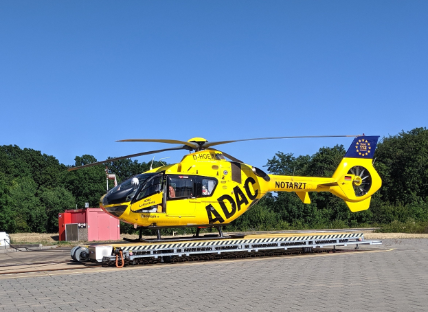 Gelber Hubschrauber auf einem Flughafenvorfeld mit Bäumen und einem klaren blauen Himmel, umgeben von Gegenständen, die wahrscheinlich mit Luftrettungsdiensten in Deutschland in Verbindung stehen.