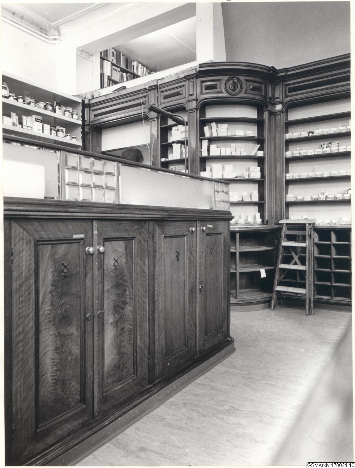 Schwarzes und weißes Foto eines vintage Apothekeninneren mit einer Theke, Schubladen, einer Leiter, Regalen voller Flaschen und Behälter und Text oben.