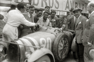 Eine Gruppe von Männern in Mützen steht um ein schwarz-weißes Rennauto herum, mit einer Tafel mit Text im Hintergrund.