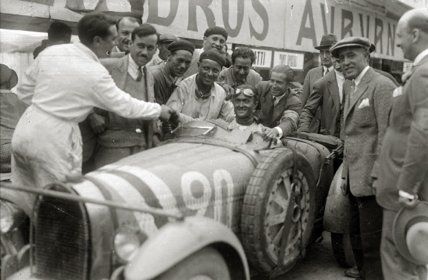Eine Gruppe von Männern in Mützen steht um ein schwarz-weißes Rennauto herum, mit einer Tafel mit Text im Hintergrund.